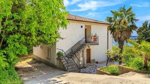 A two-story building with a staircase leading to a balcony, surrounded by greenery and a view of the sea.