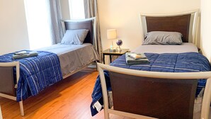 A hotel room with two single beds, a small table with a lamp, and a window with curtains.