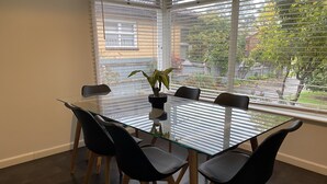 Une table à manger avec un plateau en verre, quatre chaises noires, une plante en pot et une vue sur un quartier résidentiel, visible à travers de grandes fenêtres équipées de stores.