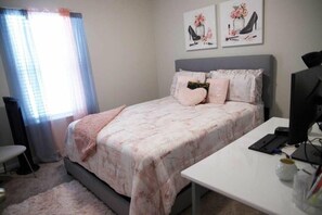 Un dormitorio con cama, escritorio y ordenador. Hay cortinas, una silla y una obra de arte en la pared.