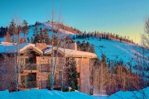 A multi-story building with a stone facade, surrounded by snow-covered ground and trees, overlooking a snowy slope. - Steps to Trails-Pool-Hot Tub-W/D in Unit-Grill (Steamboat Springs)