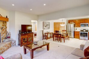 A spacious living room with a stone fireplace, a wooden coffee table, and a television. There is a dining area with wooden chairs and a chandelier. The kitchen features modern appliances and wooden cabinets. - Amazing Location on Mtn w/Air Conditioning-100 yds to Trails-Heated Pool-Hot Tub-W/D in Unit-Grill (Steamboat Springs)