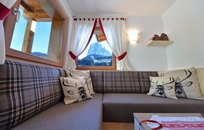 A cozy room with a sofa, a window with curtains, a wooden shelf, and a view of a mountain. - Apartment Albert with Mountain View, Wi-Fi, Balcony & Garden (St. Christina in Gröden)