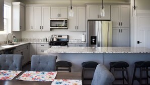 A modern kitchen with white cabinets, stainless steel appliances, and a marble countertop. There are four bar stools and a dining table with floral placemats. - Blue Lake Retreat - fully-landscaped home sits on 2nd tee of Bear Lake G.C. (Garden City)