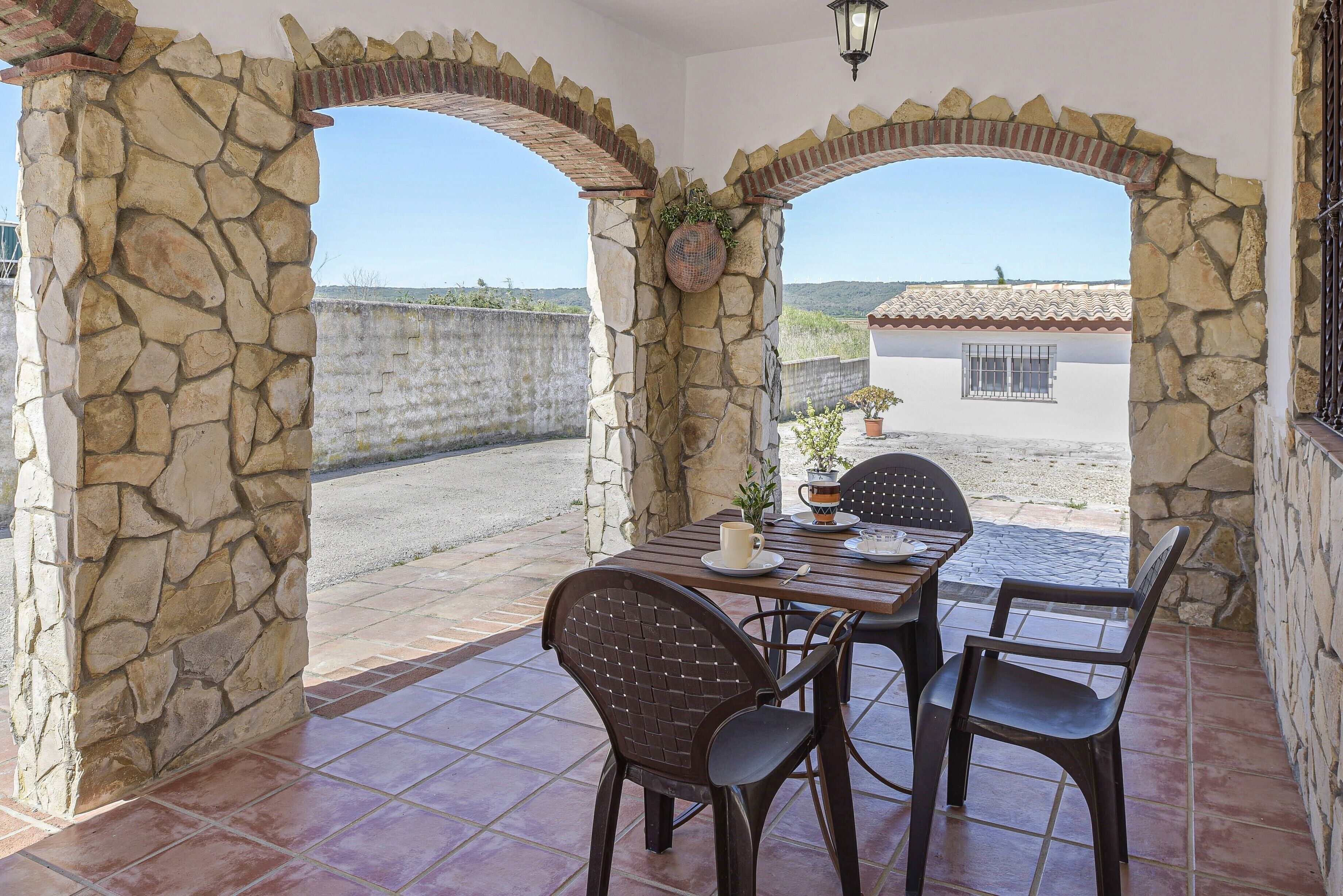 Eine terrasse mit steinbogiger Struktur, einem Tisch, Stühlen, einem Blumentopf und Blick auf ein Gebäude im Hintergrund.