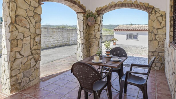 Um pátio de pedra com arco, com mesa e cadeiras, uma planta em vaso e vista para um edifício ao fundo.
