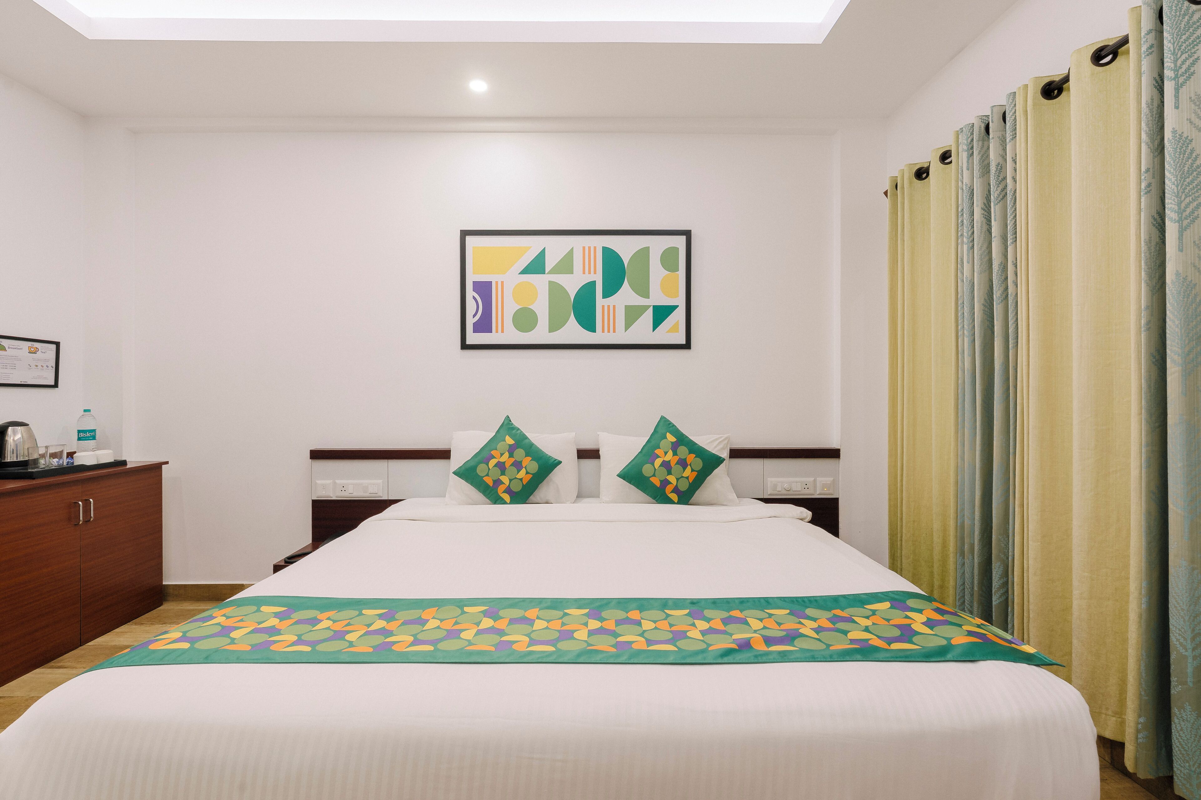 A hotel room with a large bed, a headboard with decorative pillows, a framed abstract artwork on the wall, and a window with curtains.
