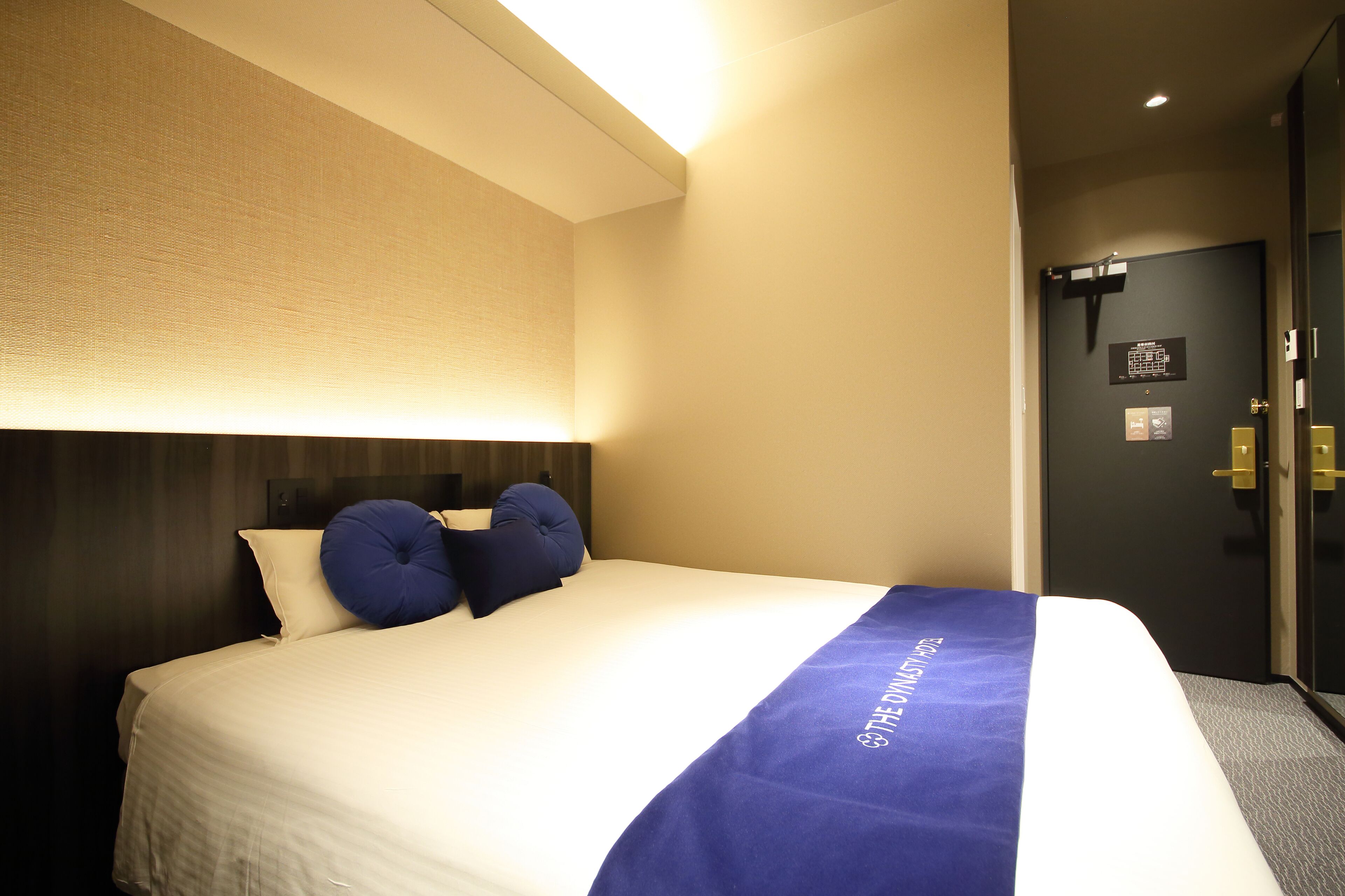 A hotel room with a neatly made bed, a headboard, and a blue accent pillow.
