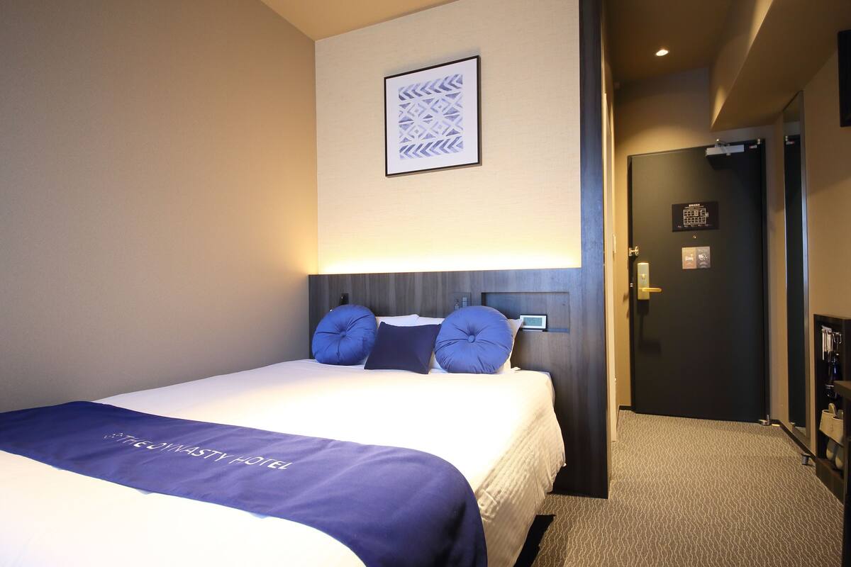 Una habitación de hotel con una cama bien hecha, un cabecero, una obra de arte enmarcada en la pared y una puerta que da a otra habitación.