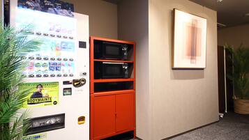 A vending machine with various snacks and drinks, a microwave, and a small orange cabinet in a hallway with a framed artwork on the wall and a potted plant.