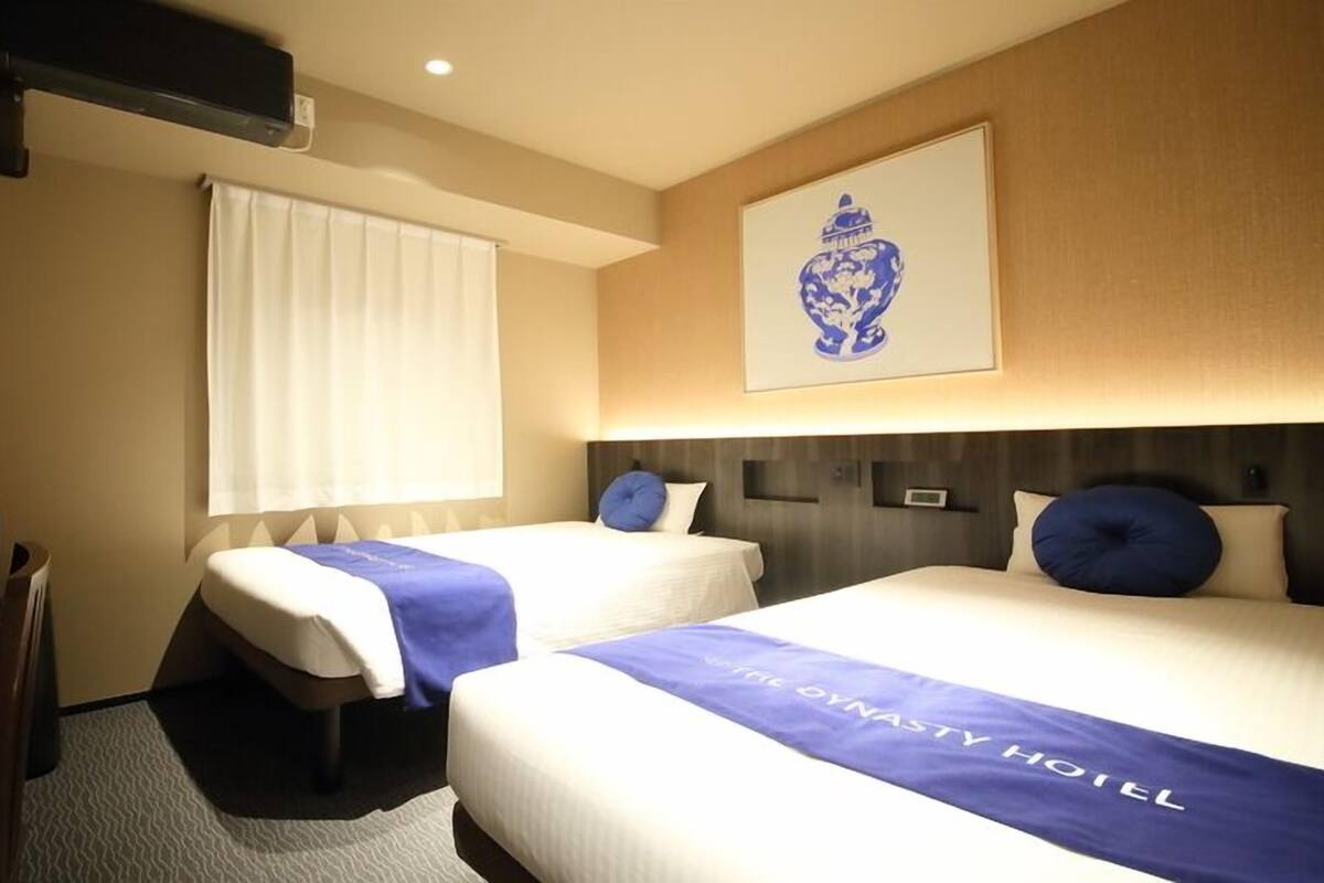 Habitación de hotel con dos camas, televisor, ventana con persianas, una obra de arte colgada en la pared y una mesita de noche.