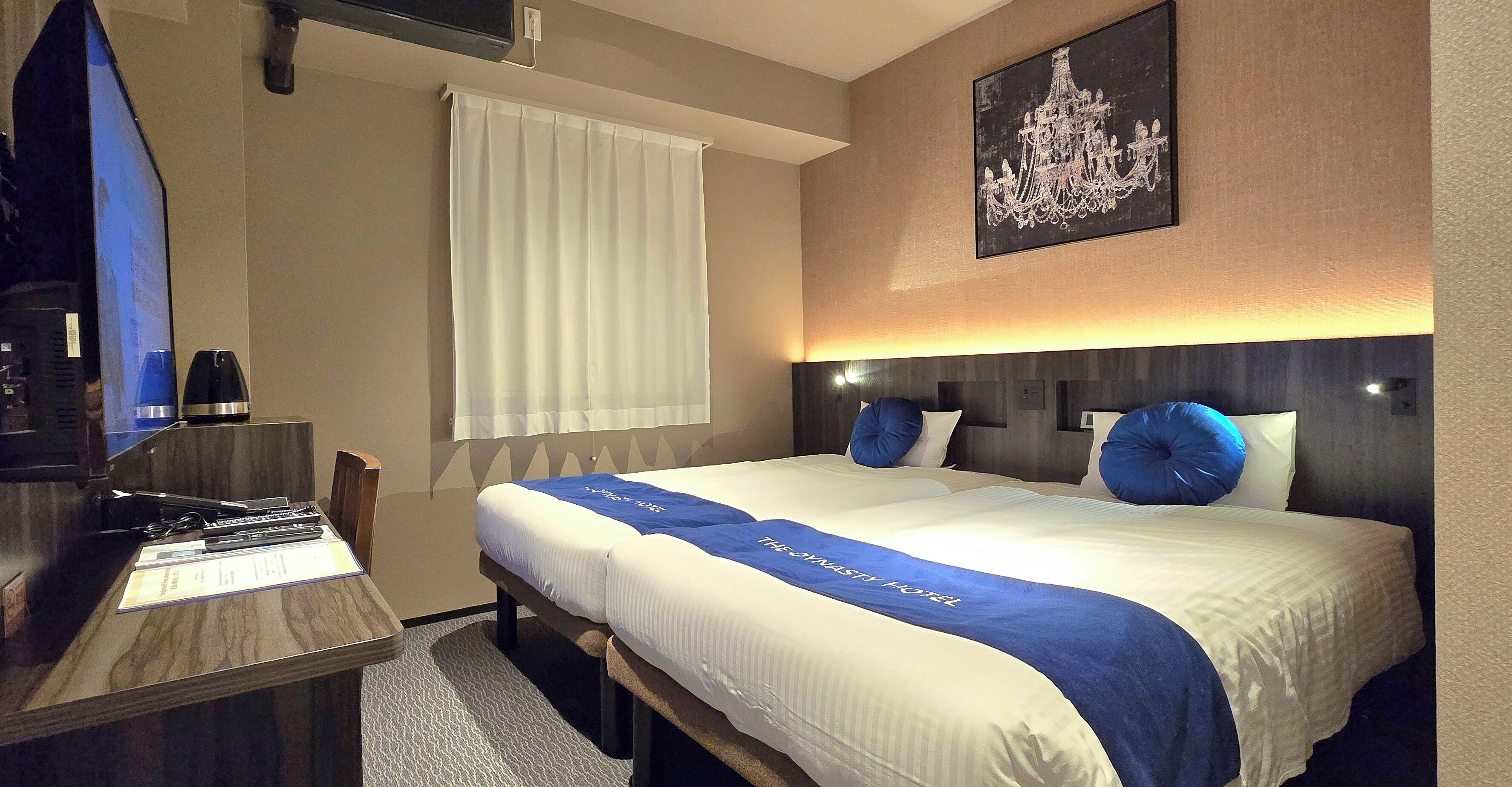 A hotel room with a bed, a desk with a computer, a chair, a window with curtains, and a chandelier on the wall.