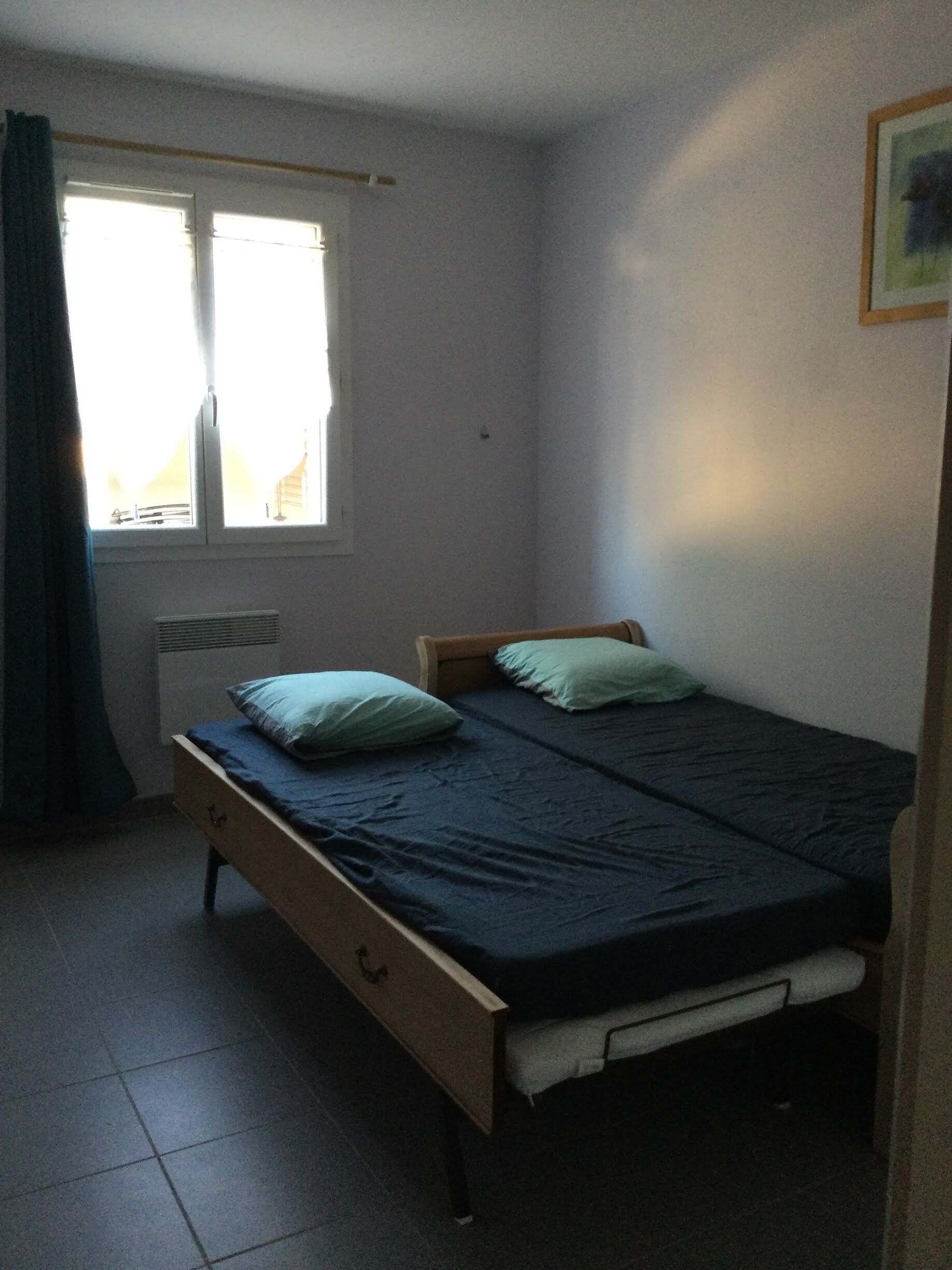Quarto pequeno com cama de solteiro, janela, radiador e estrutura de cama em madeira.