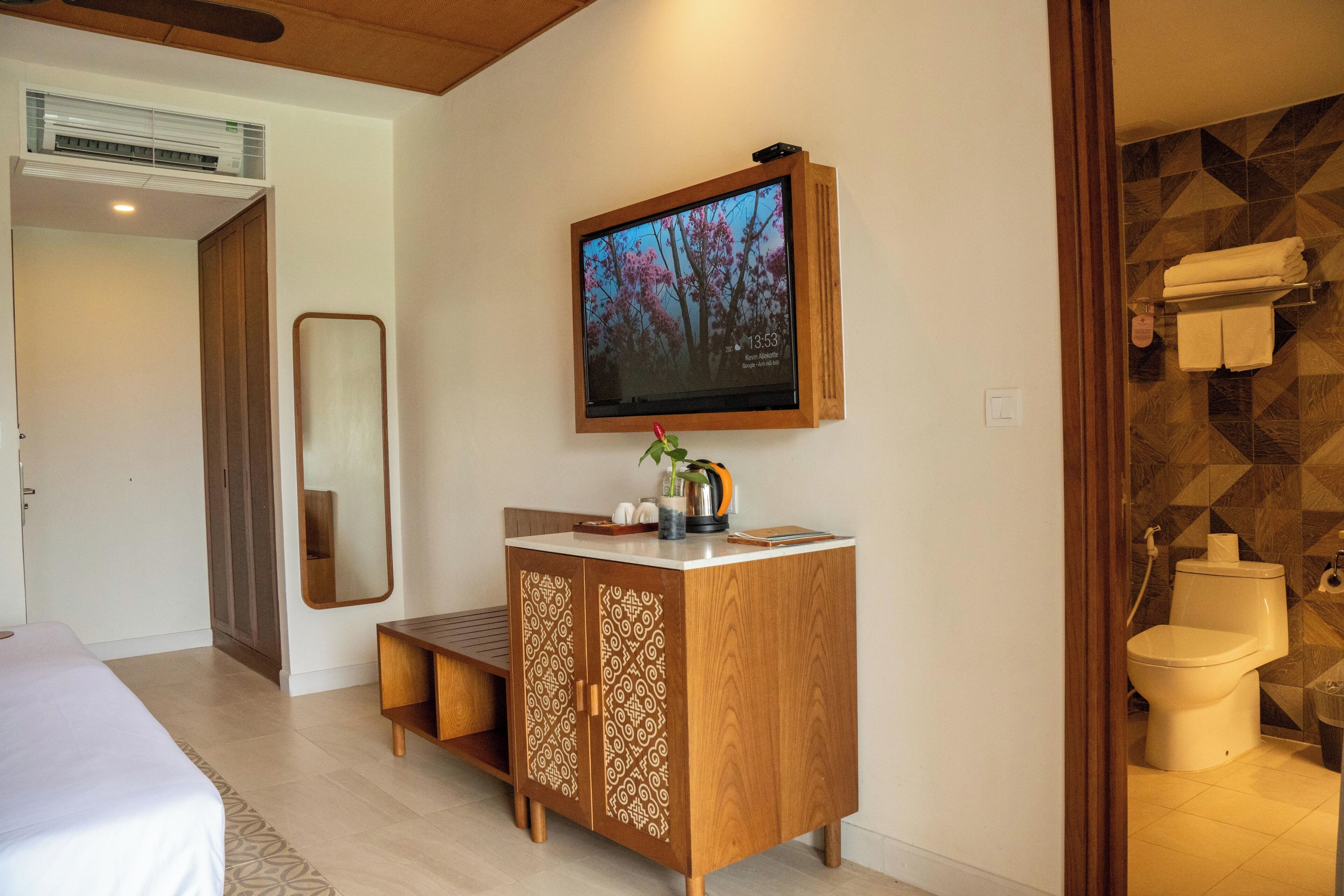A hotel room with a bed, a wooden cabinet with decorative patterns, a mounted TV, and a bathroom with a toilet and towel rack.