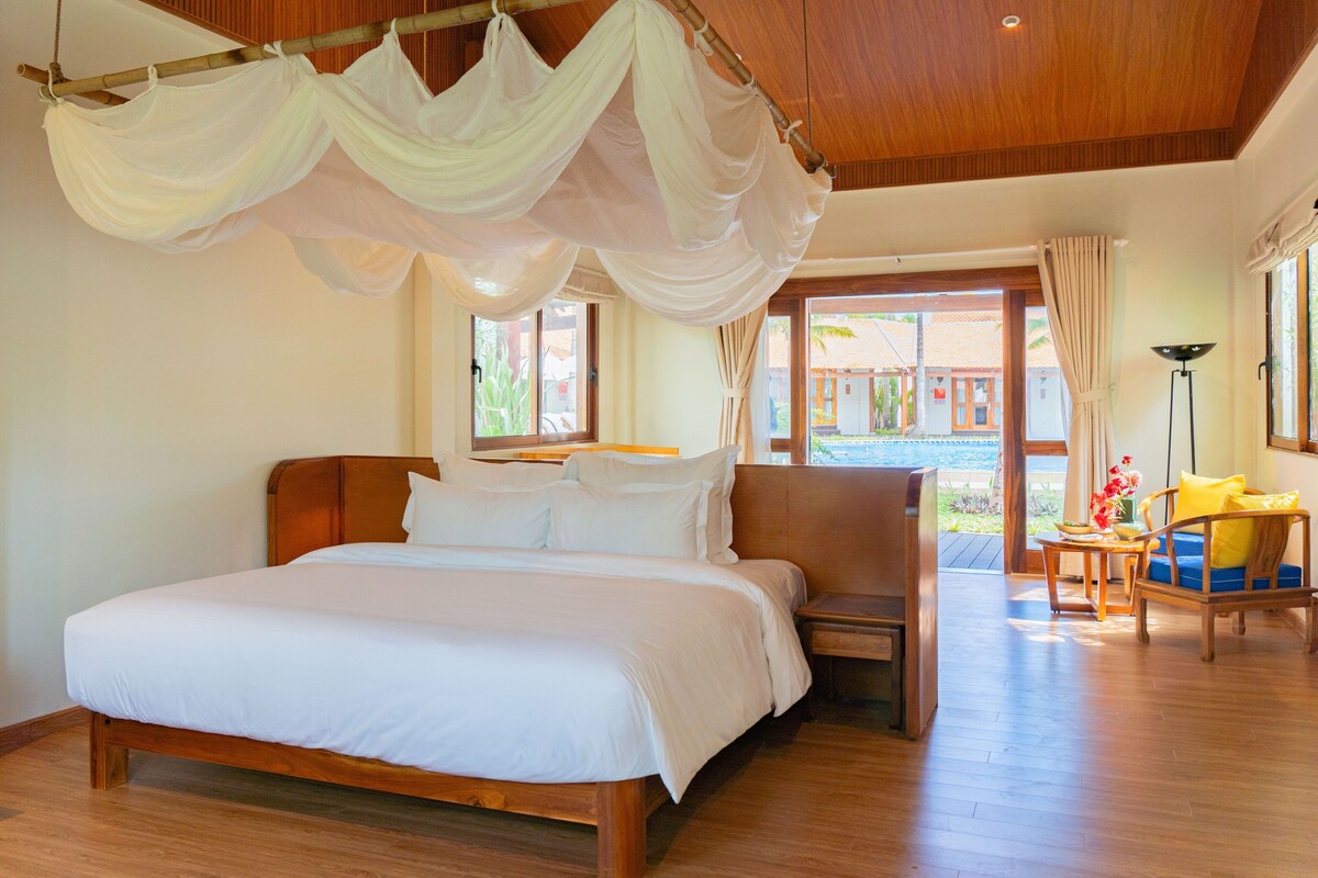 Un dormitorio con una cama grande, cabecera de madera, mesita de noche, una silla y vistas al exterior.