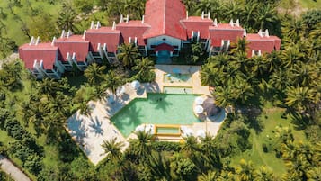 Vista aérea de um resort com vários edifícios de telhado vermelho, uma piscina central e palmeiras ao redor.