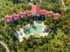 An aerial view of a resort with multiple red-roofed buildings, a central swimming pool, and surrounding palm trees. - Pax Ana Doc Let Resort & Spa (Ninh Hoa)