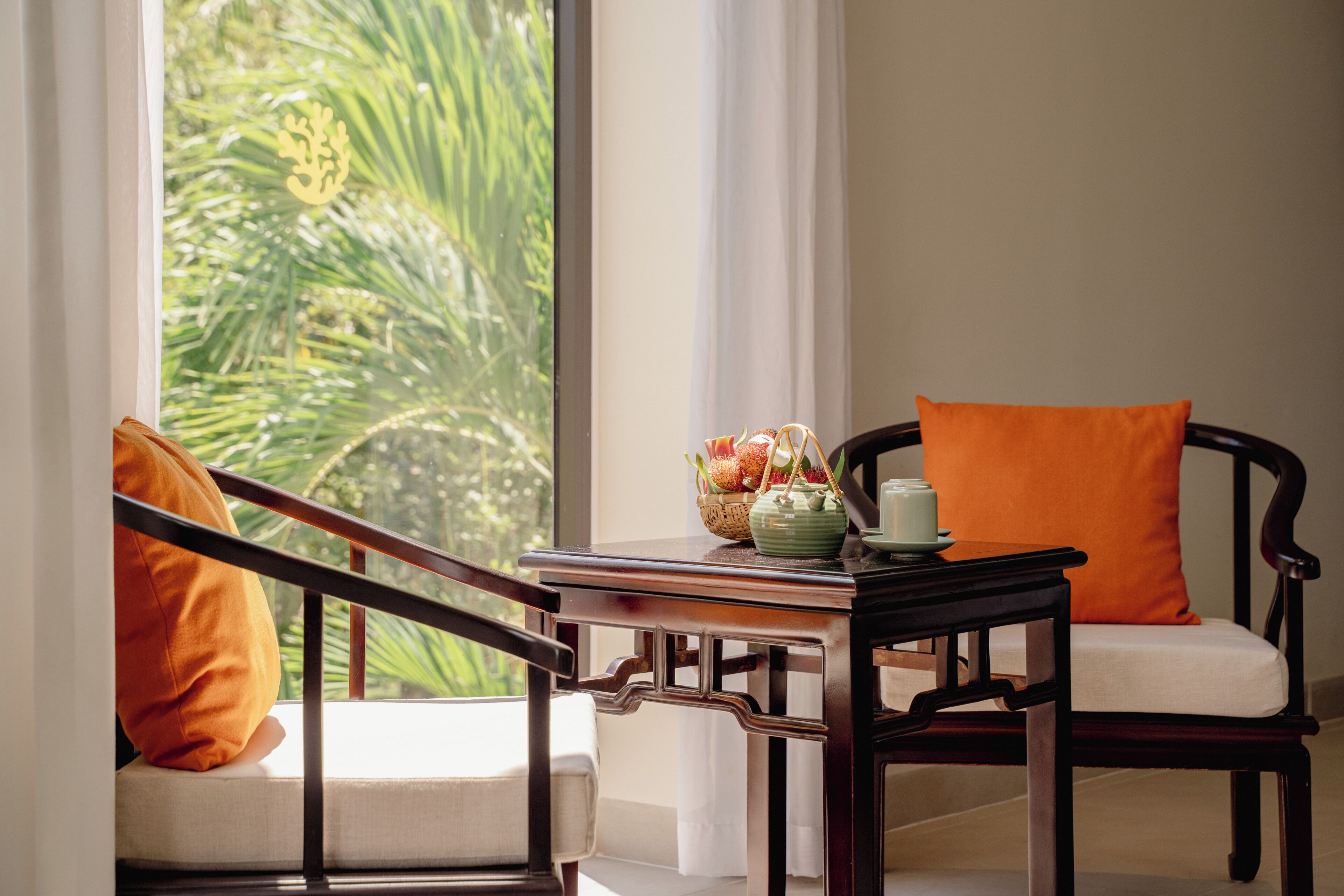Una habitación con vistas a la vegetación a través de una ventana, una mesa con tetera y fruta, y una silla con cojín naranja.