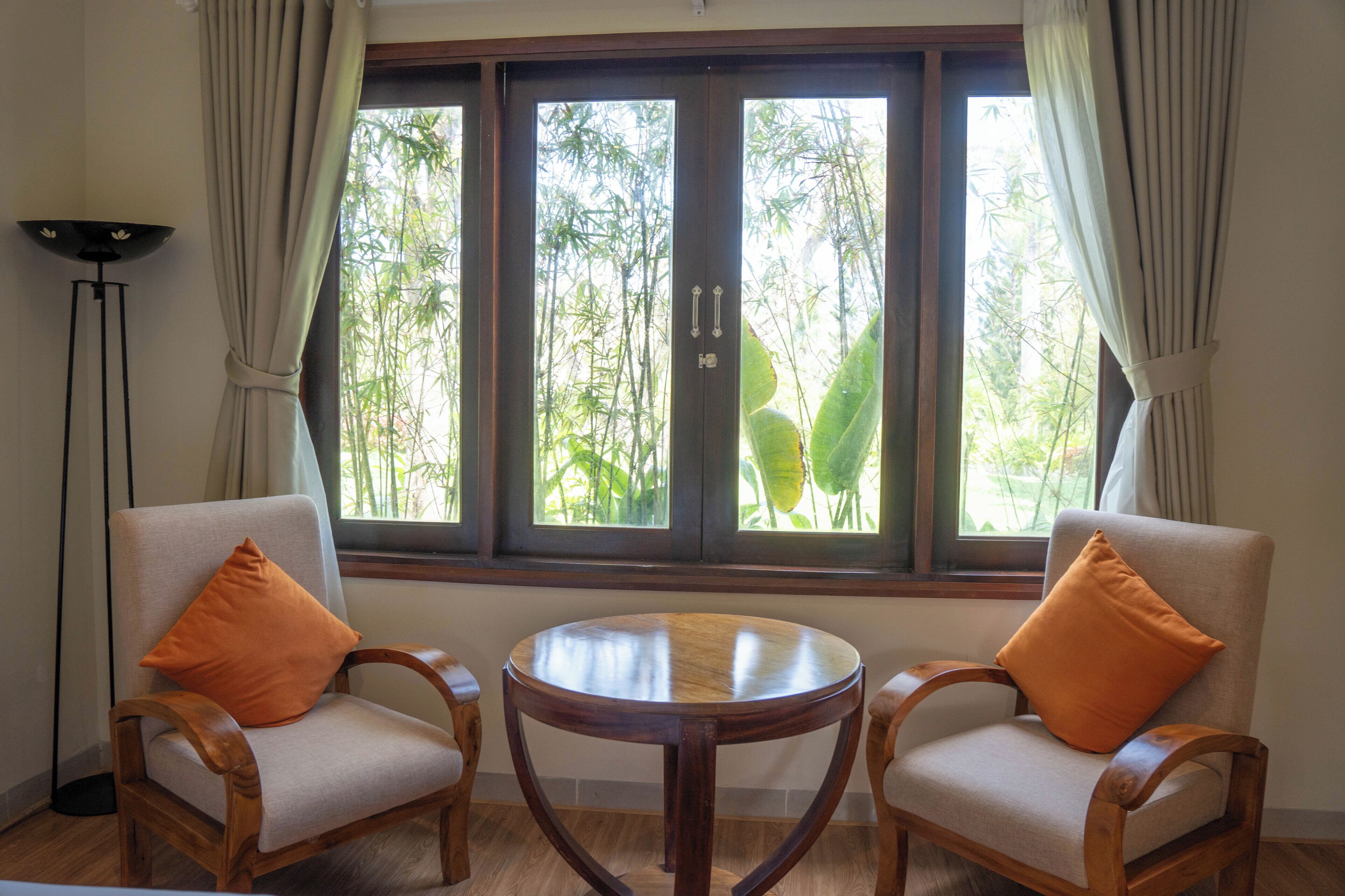 Una habitación con dos sillas de madera con cojines, una mesita de madera y un ventanal con cortinas.
