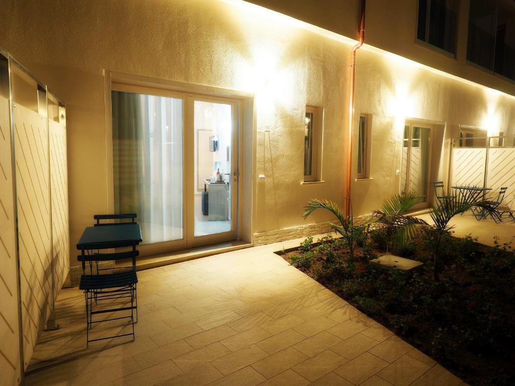 Un patio esterno ben illuminato con tavolo e sedie, un piccolo giardino con piante e una porta scorrevole in vetro che dà su un ambiente interno.