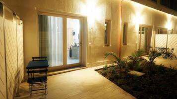 Un patio esterno ben illuminato con tavolo e sedie, un piccolo giardino con piante e una porta scorrevole in vetro che dà su un ambiente interno.