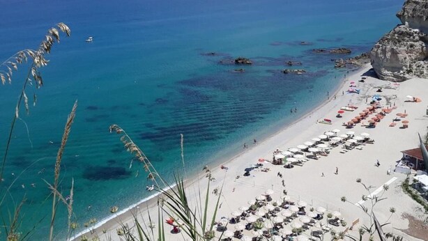 A beach with clear blue water, a rocky cliff, and a row of beach umbrellas and lounge chairs. - Tui Blue Tropea – Baia di Riaci Resort (Santa Domenica)