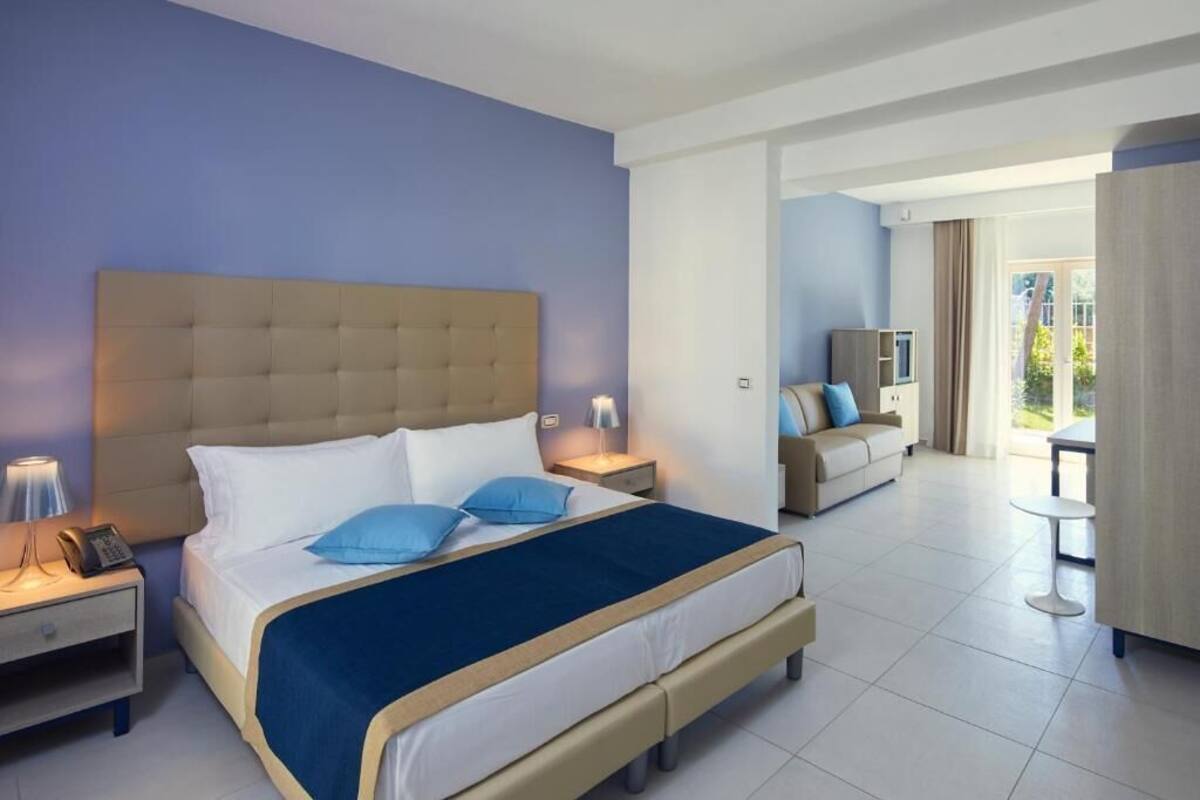 A hotel room with a large bed, a bedside table with a lamp, a nightstand, a sofa, and a view of the outdoors.