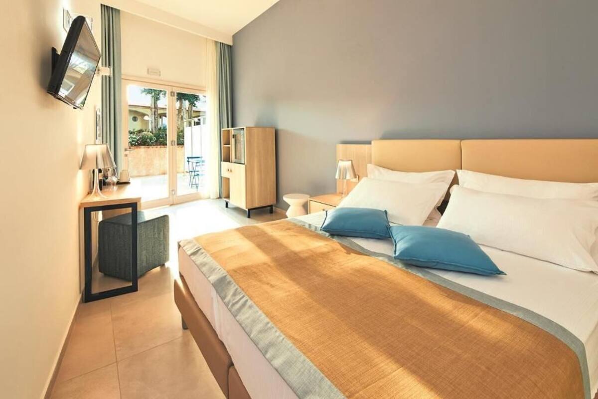 A hotel room with a large bed, a TV mounted on the wall, a desk with a lamp, and a view of the outside.
