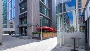 A modern building with a glass facade, outdoor seating area with red umbrellas, and a small table.