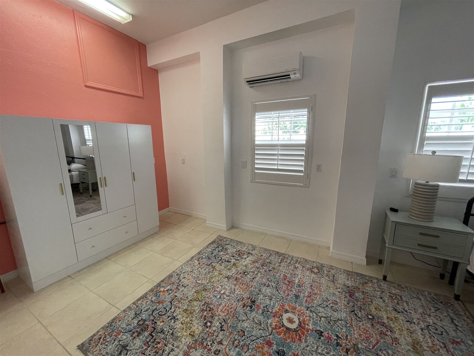 A room with a large wardrobe, a patterned rug, a window with blinds, and an air conditioning unit.
