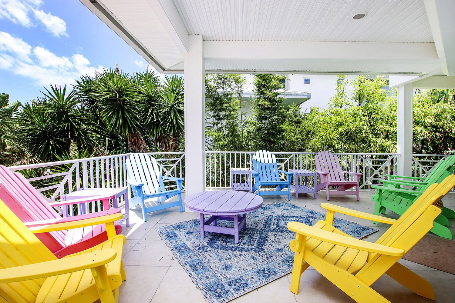 Une terrasse couverte, agrémentée de chaises Adirondack aux couleurs vives et d’une petite table, le tout entouré d’une balustrade blanche et d’une végétation luxuriante.