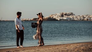 Escena de playa con una persona con vestido estampado y sombrero, y otra persona con camisa blanca.