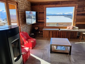 Ein gemütliches Blockhaus-Interieur mit Steinwand, Holzbalkendecke, Wand-Fernseher, rotem Ledersessel, einer Holzbank und einem kleinen Tisch.