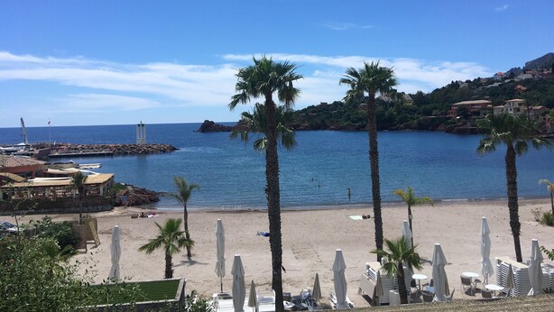 A beach with palm trees, a pier, and buildings on a hillside. - Séjour de Rêve sur la Côte D'azur (Théoule-sur-Mer)