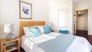 Una camera da letto ordinata con una testiera in legno, un letto con biancheria azzurra e bianca, una lampada da comodino, un quadro appeso al muro e una porta che dà su un'altra stanza.