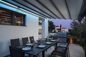 An outdoor dining area with a table set for four, a grill, and a pool in the background.