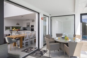 Een moderne keuken met witte kasten, een eethoek met een ronde tafel en rieten stoelen, en een glazen deur naar een terras met uitzicht.