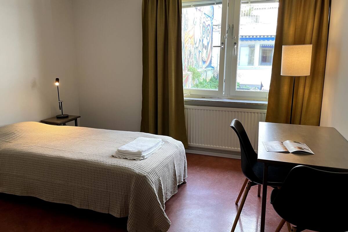 A hotel room with a bed, a desk, a chair, a lamp, and a window with curtains.
