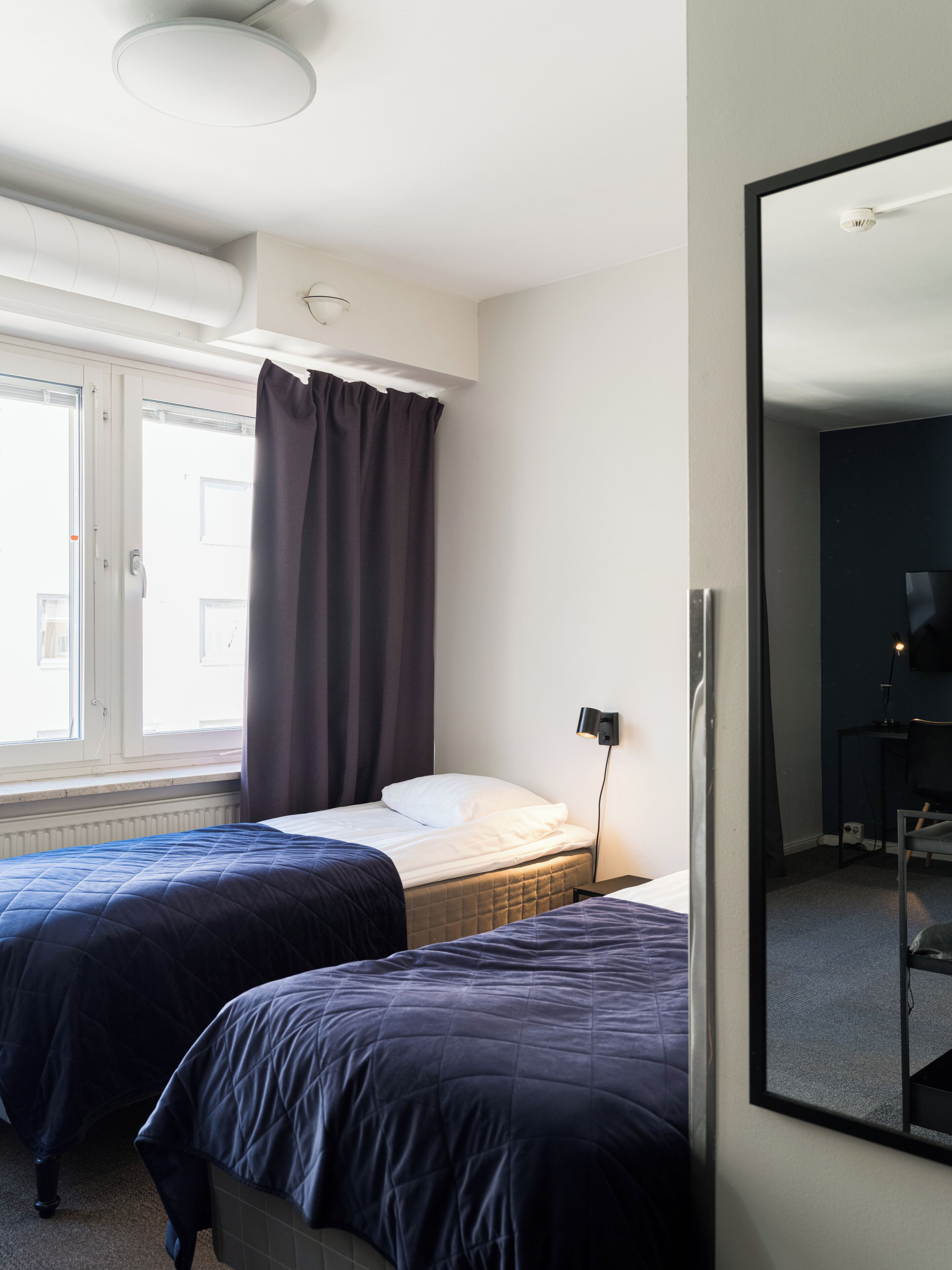 Quarto de hotel com duas camas, um espelho grande, uma escrivaninha e uma janela com cortinas.