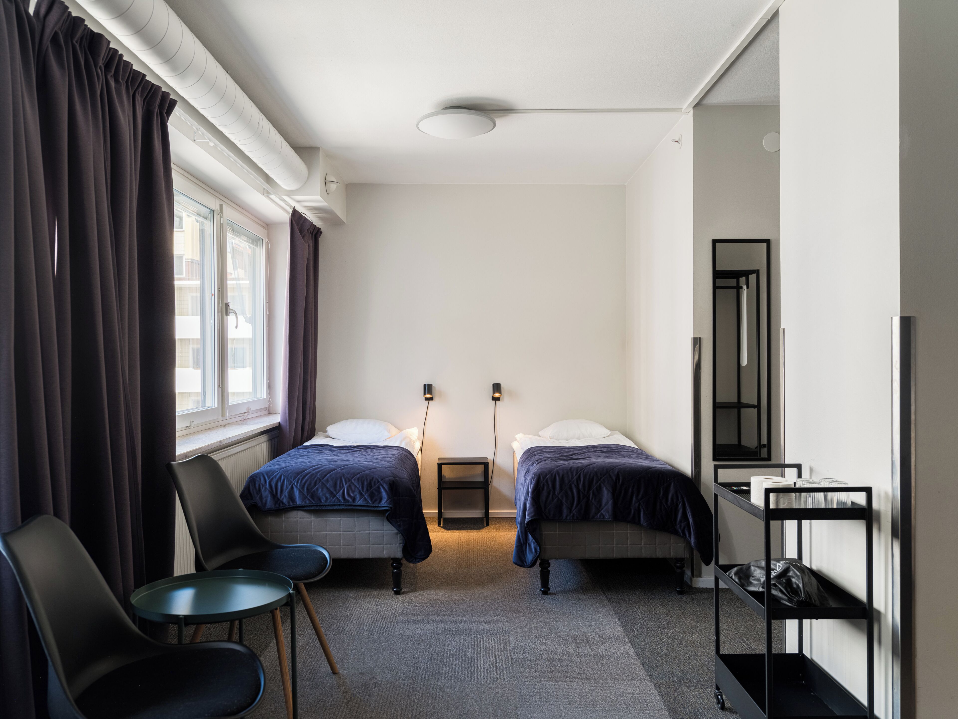 A hotel room with two beds, a chair, a small table, a radiator, a mirror, and a shelf with items.
