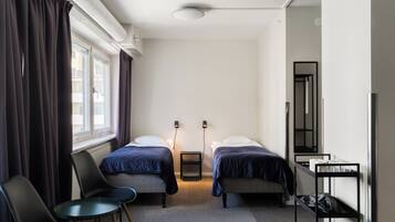 A hotel room with two beds, a chair, a small table, a radiator, a mirror, and a shelf with items.