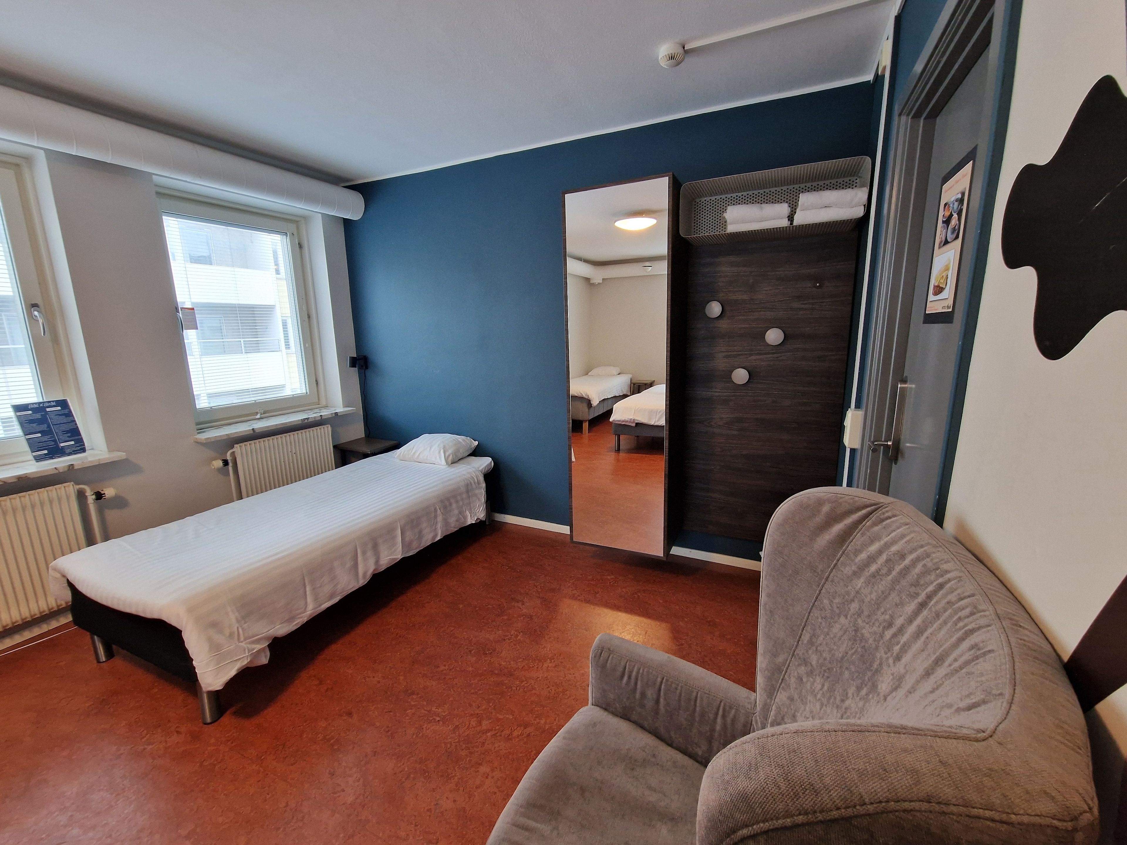 Habitación individual con cama, ventana, radiador, espejo y sillón gris.