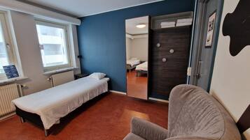 Habitación individual con cama, ventana, radiador, espejo y sillón gris.
