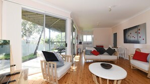 Un vaste espace de vie avec une grande porte-fenêtre coulissante donnant sur un jardin, un canapé blanc avec des coussins, une table basse et un tableau accroché au mur.