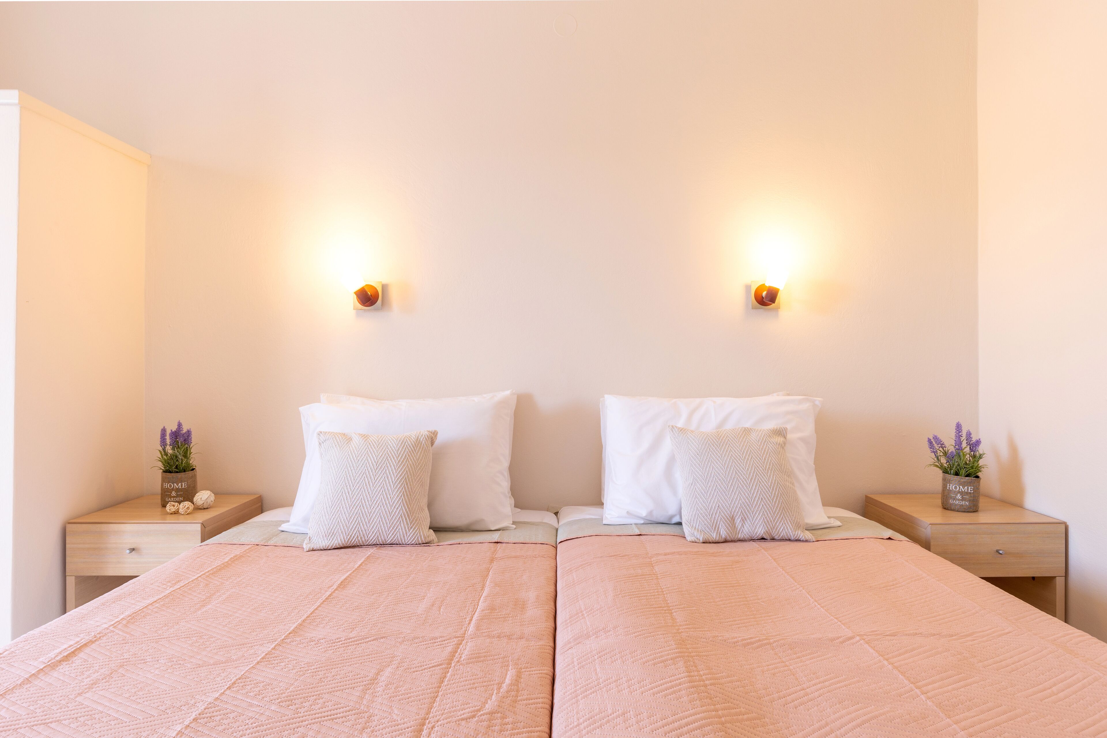 A double bed with a pink quilt, two white pillows, and a small plant on a bedside table.