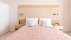Quarto bem arrumado com cabeceira de madeira, cama com colcha rosa, duas mesinhas de cabeceira com objetos decorativos e um guarda-roupa branco.