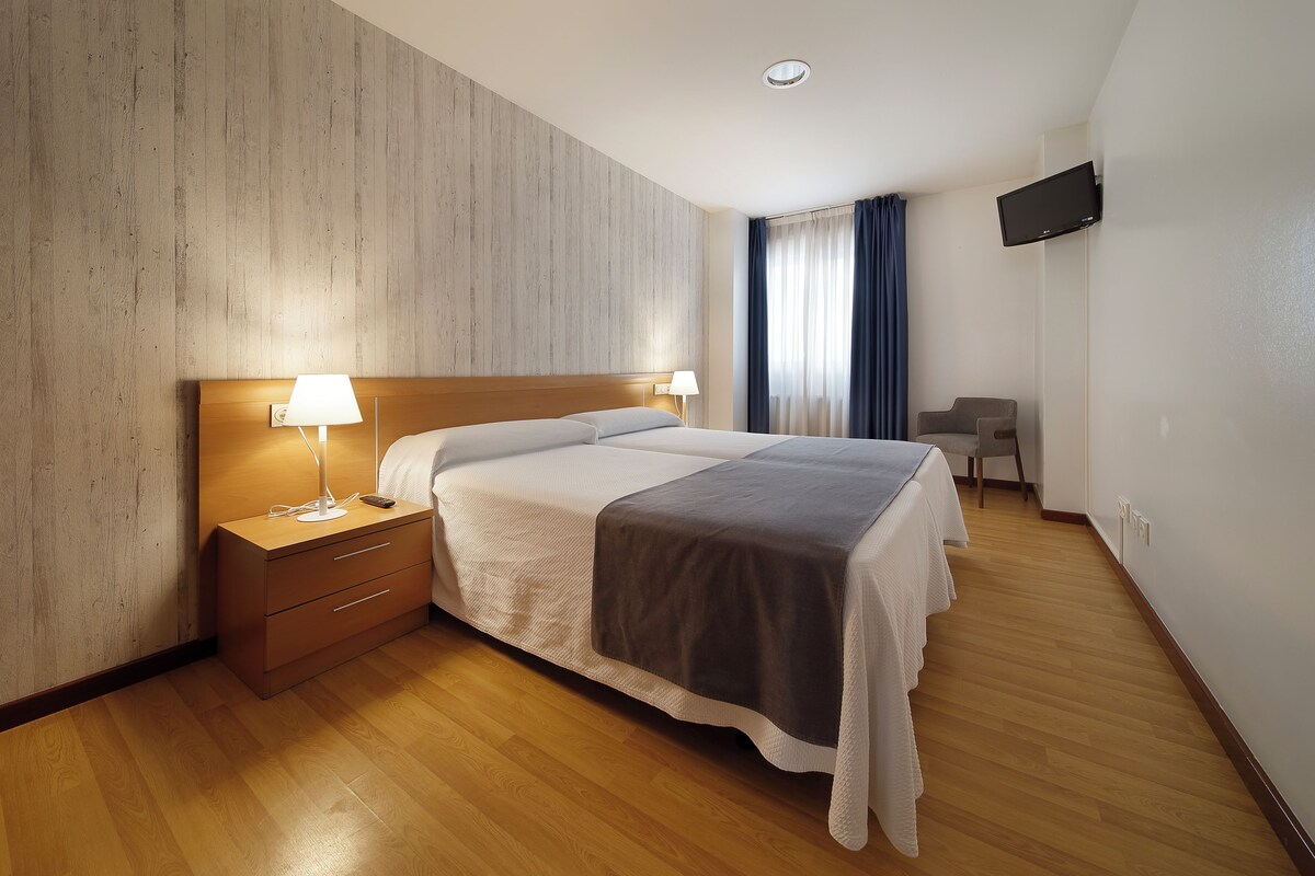 A hotel room with a wooden headboard bed, a bedside table with a lamp, a TV mounted on the wall, and a small seating area with a chair and table.
