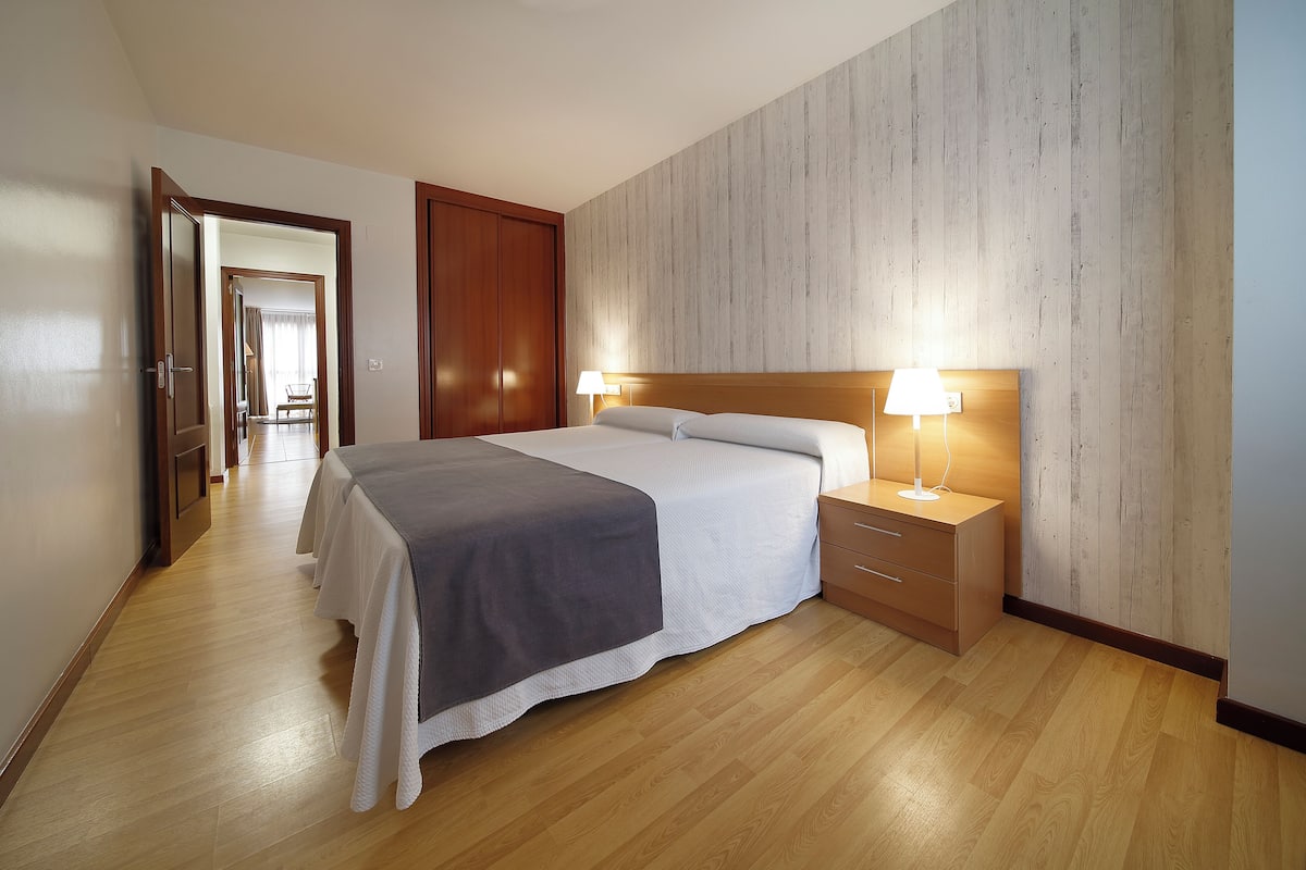 Ein Hotelzimmer mit Holzboden, einem Bett mit grau-weißer Tagesdecke, einem Nachttisch aus Holz mit einer Lampe und einer Holztür.