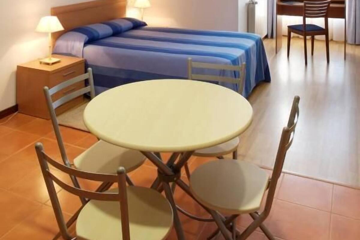 Ein kleiner, gemütlicher Raum mit einem Bett, einem runden Tisch und zwei Stühlen. Es gibt einen Nachttisch mit einer Lampe und einem Sessel.