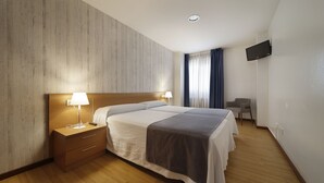 Ein Hotelzimmer mit einem Bett mit Holz-Kopfteil, einem Nachttisch mit Lampe, einem an der Wand befestigten Fernseher und einer kleinen Sitzecke mit Stuhl und Tisch.